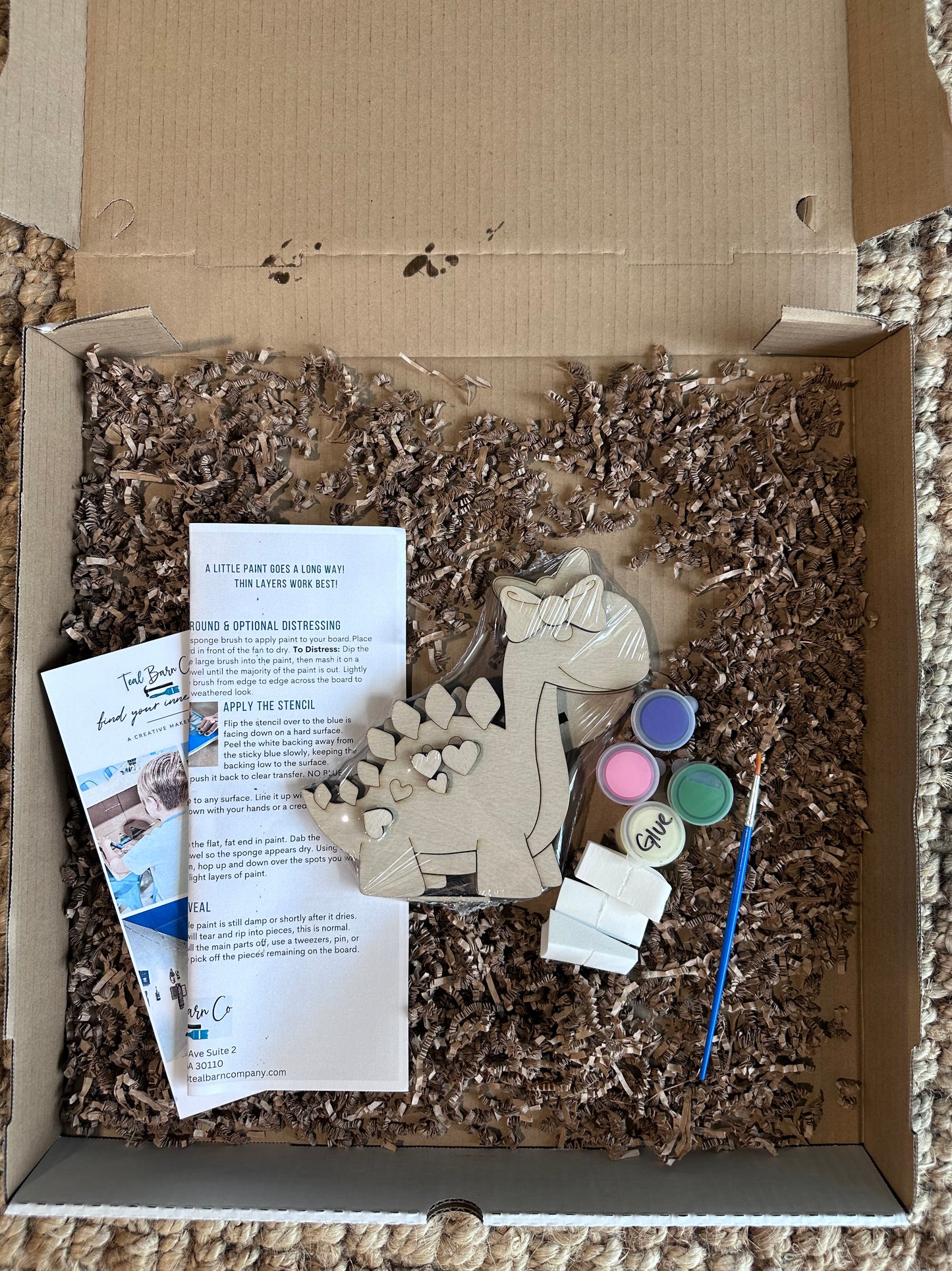 Box filled with craft materials including a wooden dinosaur figure, paint pots, and directions.