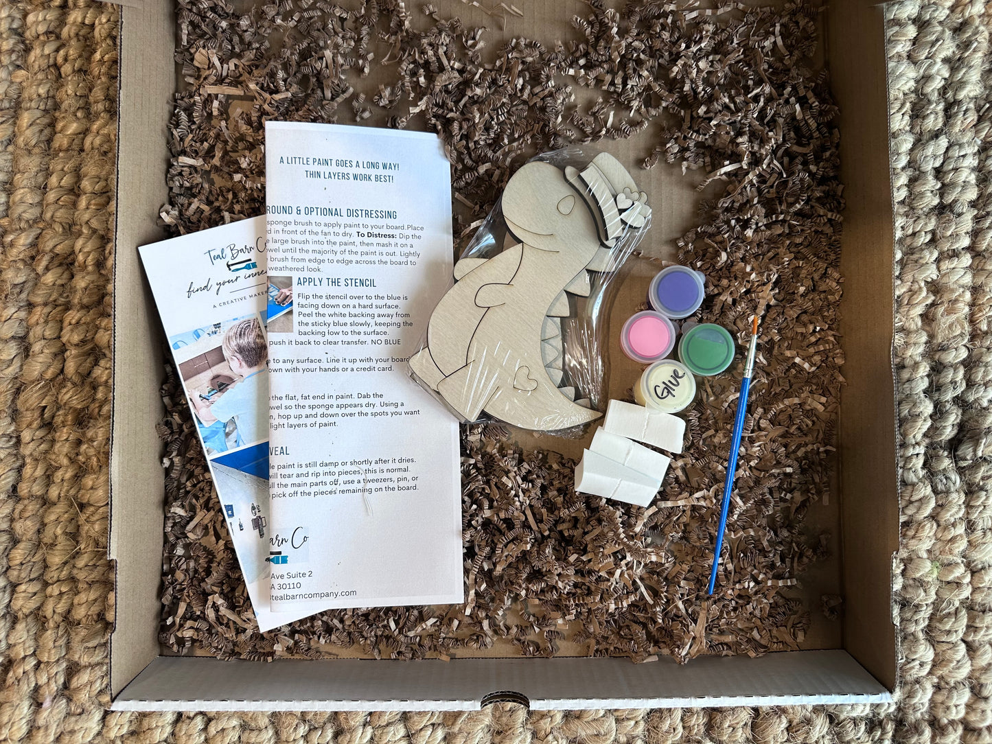 Craft kit with wooden dinosaur figure with hat, paint, brush, and instructions in a box.