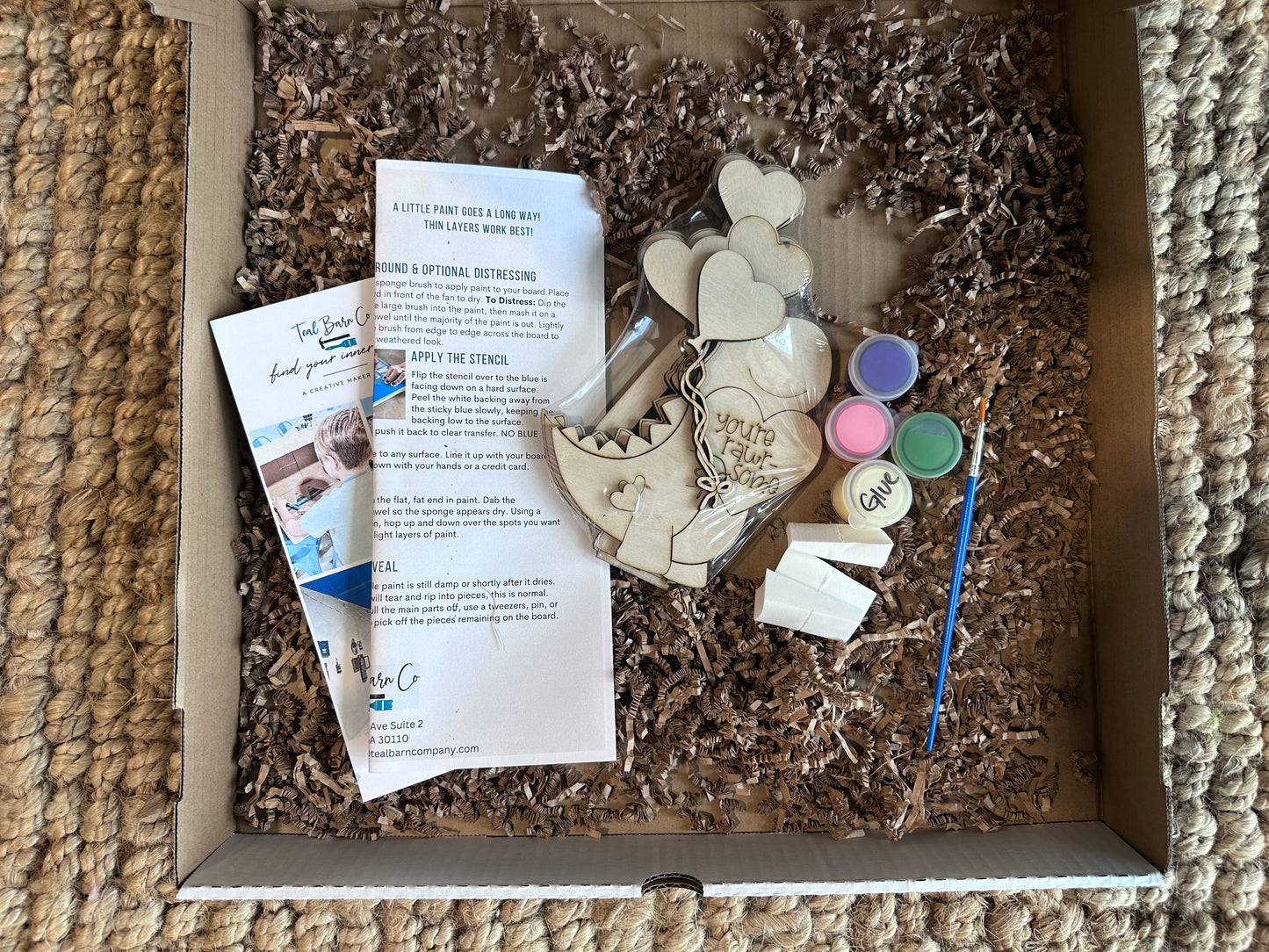 Crafting kit with dinosaur holding balloons, paint, and instructions in a cardboard box.