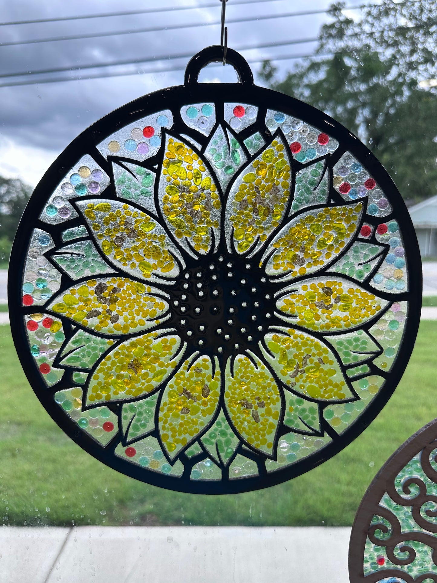 Decorative sunflower-shaped window hanging with colorful mosaic design. The background is multicolored, the petals are yellow, and the leaves are green.