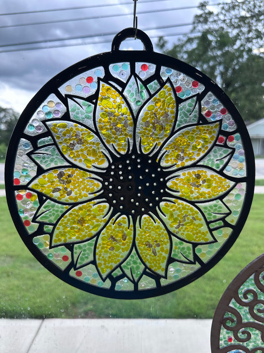 Decorative sunflower-shaped window hanging with colorful mosaic design. The background is multicolored, the petals are yellow, and the leaves are green.