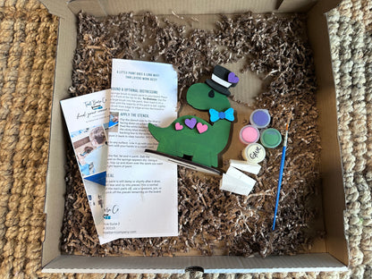 Box with craft materials including a dinosaur-shaped wooden laser cut piece, paint, and tools.