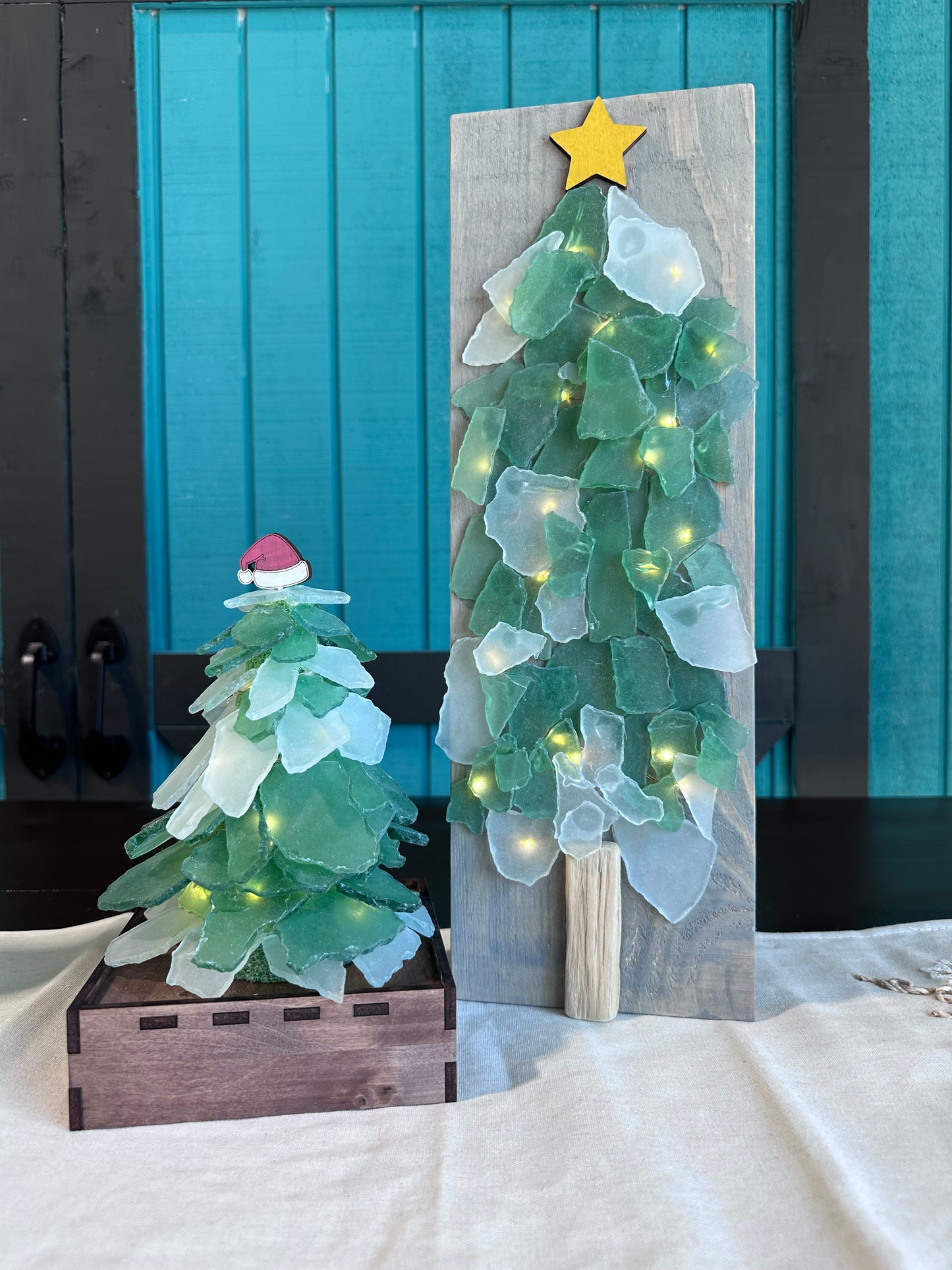 Two sea glass tree options shown: 3-D on a wooden box base, or flat on a wooden plank. Both made with various shades of green glass, and a Santa hat/star tree topper.
