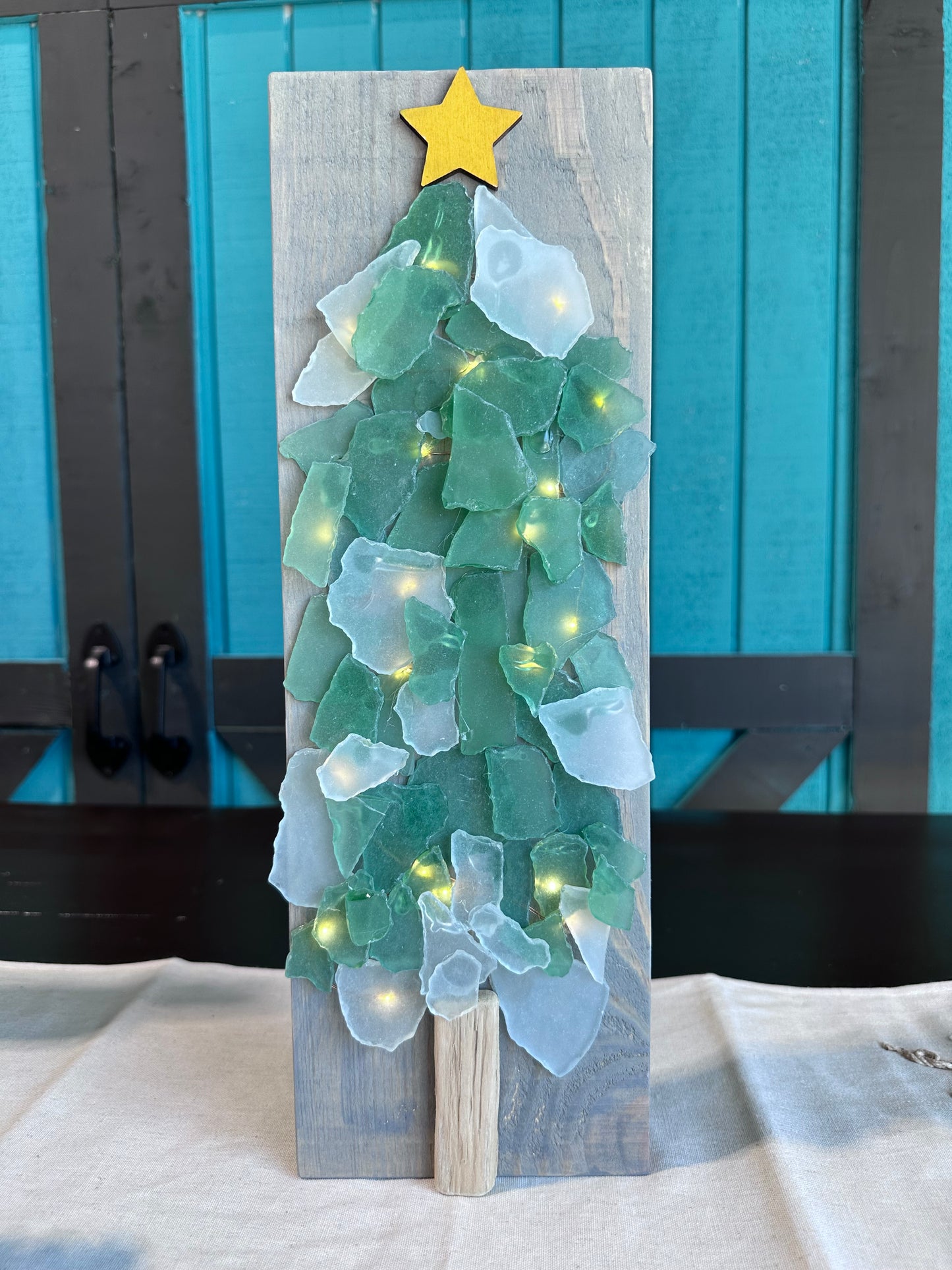 A wooden plank is shown, with green sea glass arranged on it in a Christmas tree shape. Lights are behind it to give a glow, and it is topped with a star.