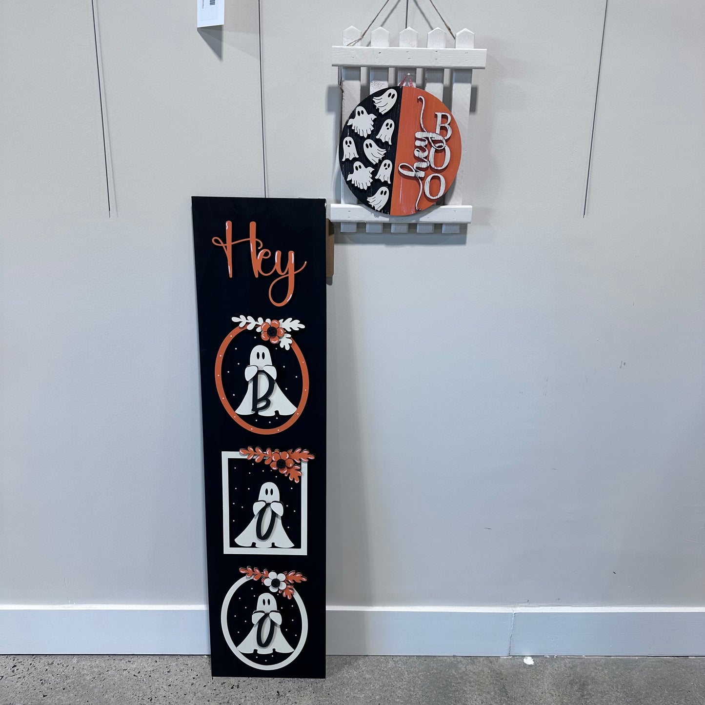 Halloween-themed decorative signs. One is a porch-leaner saying "hey boo" with three frames holding three ghosts. The other is a smaller hanging sign on a picket fence background saying "hey boo" with ghosts off to the side.