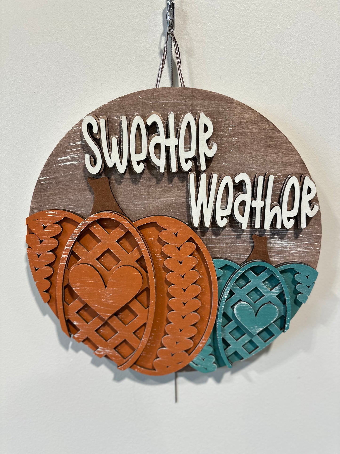 circular fall sign with two pumpkins on it, orange and teal. pumpkins have a knit-like design.