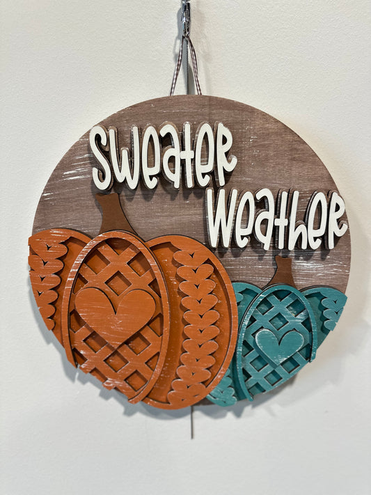 circular fall sign with two pumpkins on it, orange and teal. pumpkins have a knit-like design.