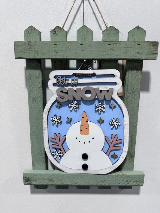 Decorative sign on picket fence. Mason jar shape, wintery design with snowman catching snowflakes. Sign is interchangeable with others on the fence.
