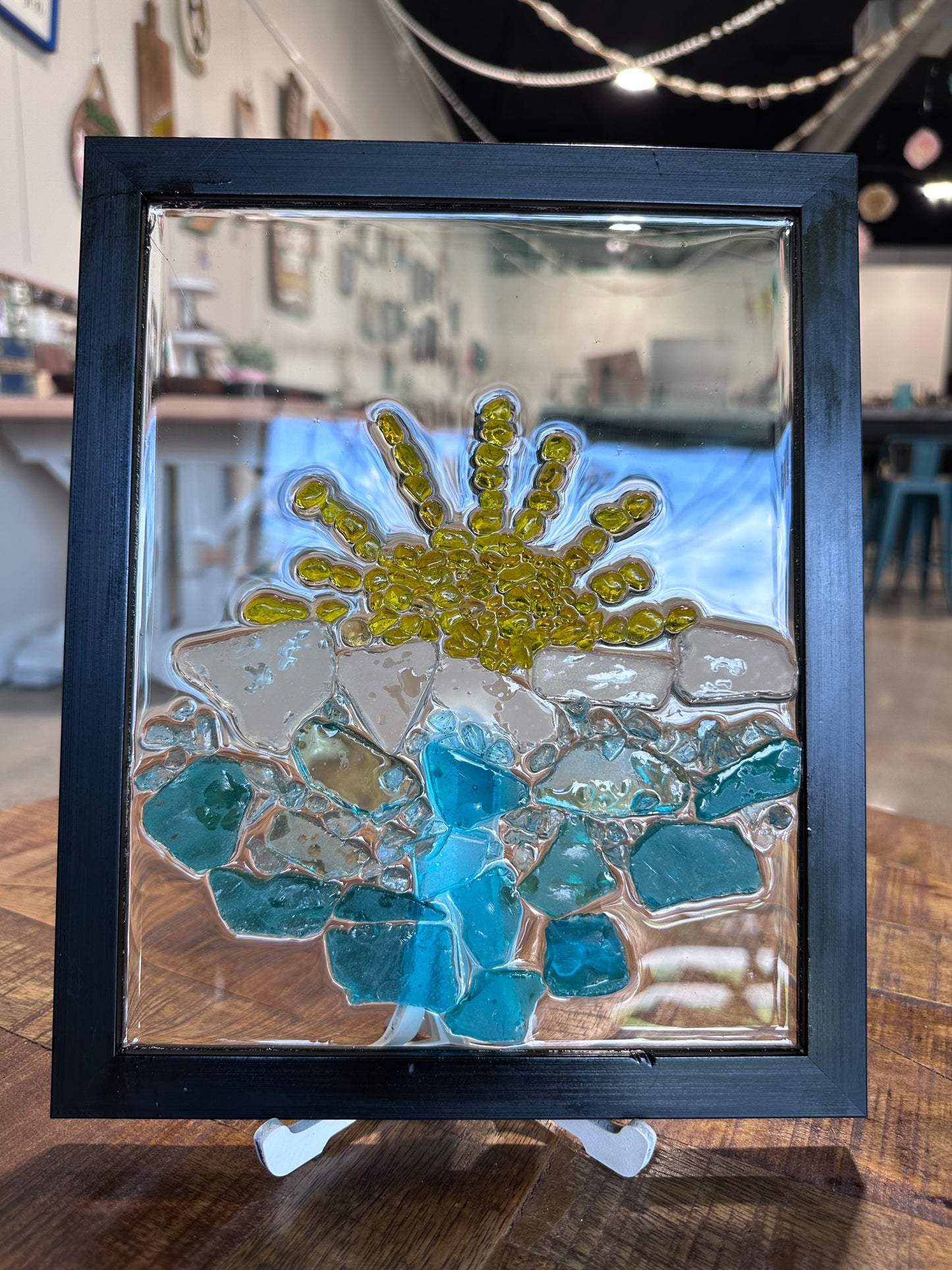 Framed glass mosaic with blue and yellow beach design on a wooden surface. The waves are blue sea glass and the sun is small yellow stones.