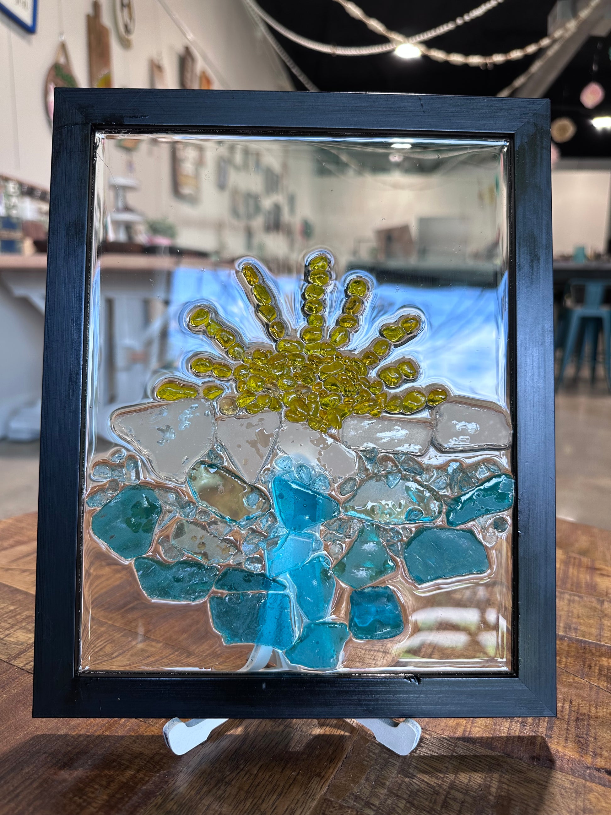 Framed glass mosaic with blue and yellow beach design on a wooden surface. The waves are blue sea glass and the sun is small yellow stones.