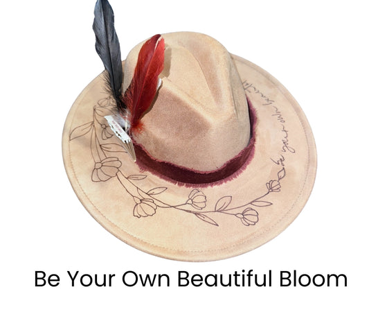 A beige hat with a floral pyrography design and a red band, with a feather embellishment on the side.